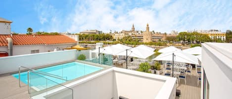 A rooftop pool with glass railings, outdoor seating, and a view of historic buildings in the background.