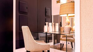 Een moderne kamer met een glazen bureau, een beige stoel en een grote spiegel die een lamp en vazen weerspiegelt.