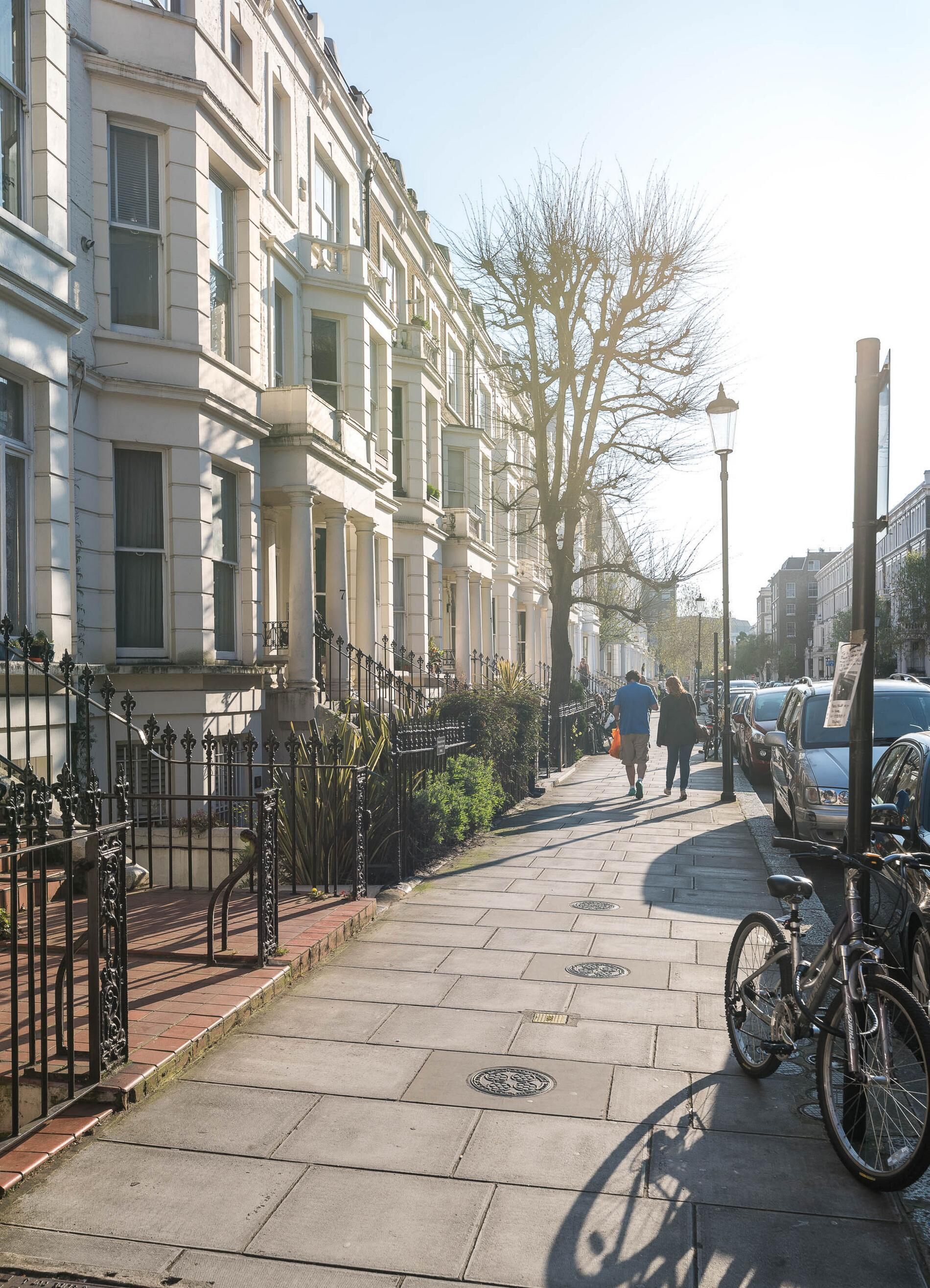 Un trottoir pavé, avec un vélo garé sur le côté droit, bordé d’immeubles dotés de balcons et d’un arbre en arrière-plan.