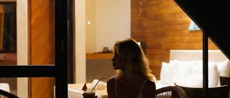 A person sitting at a table with a drink, in a room with a wooden ceiling and a bed in the background.
