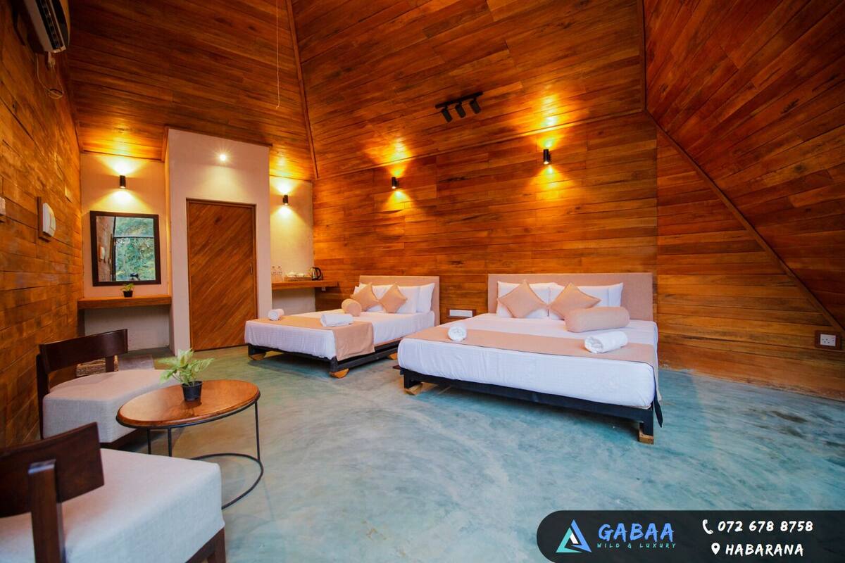 A room with two beds, a small table, and a chair. There is a window with a view of greenery. The room has wooden walls and a sloped ceiling.