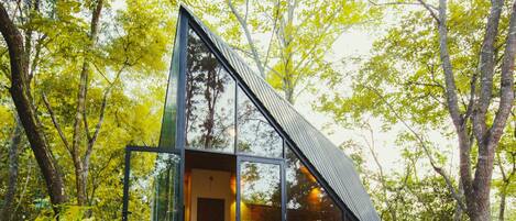 Una cabaña moderna con paredes de vidrio en un bosque, con una cama y zona de estar a la vista.