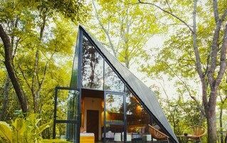 A modern, glass-walled cabin in a forest, with a visible bed and seating area.