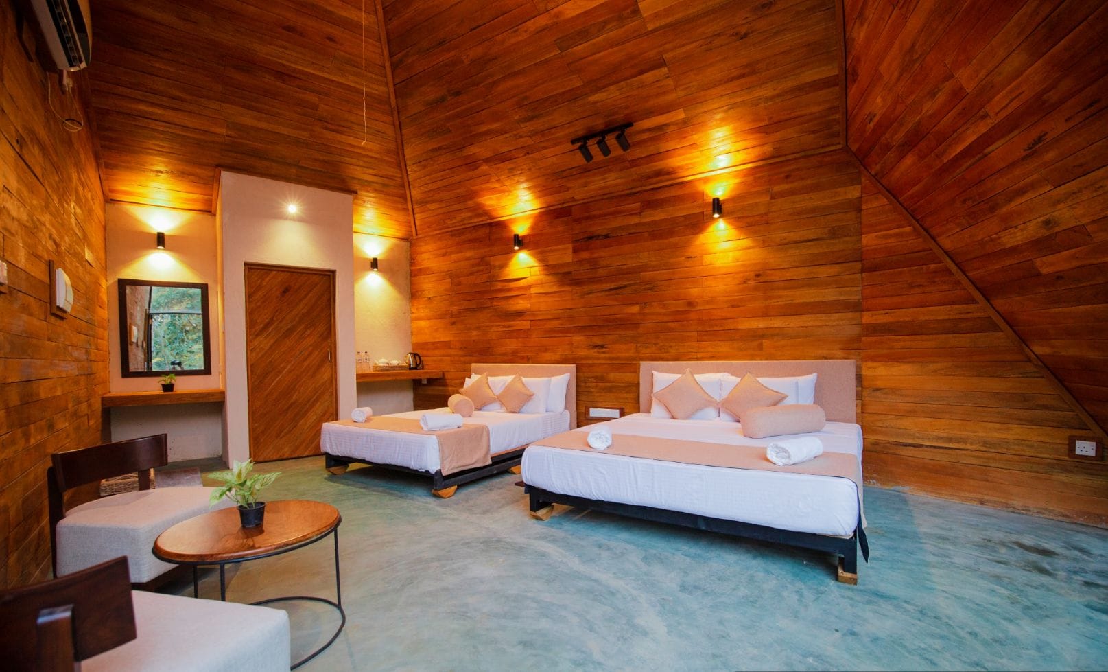A room with two beds, a small table, and a chair. There is a window with a view of greenery. The room has wooden walls and a sloped ceiling.