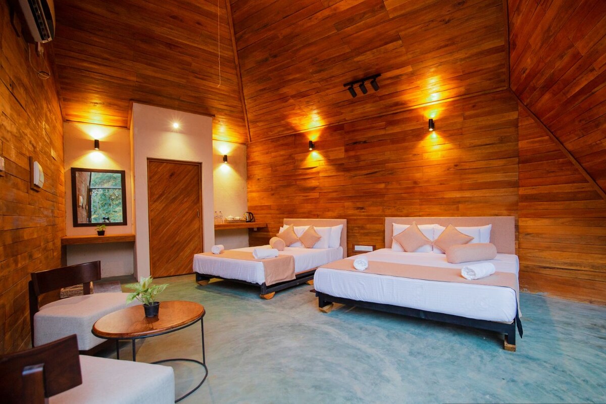 A room with two beds, a small table, and a chair. There is a window with a view of greenery. The room has wooden walls and a sloped ceiling.