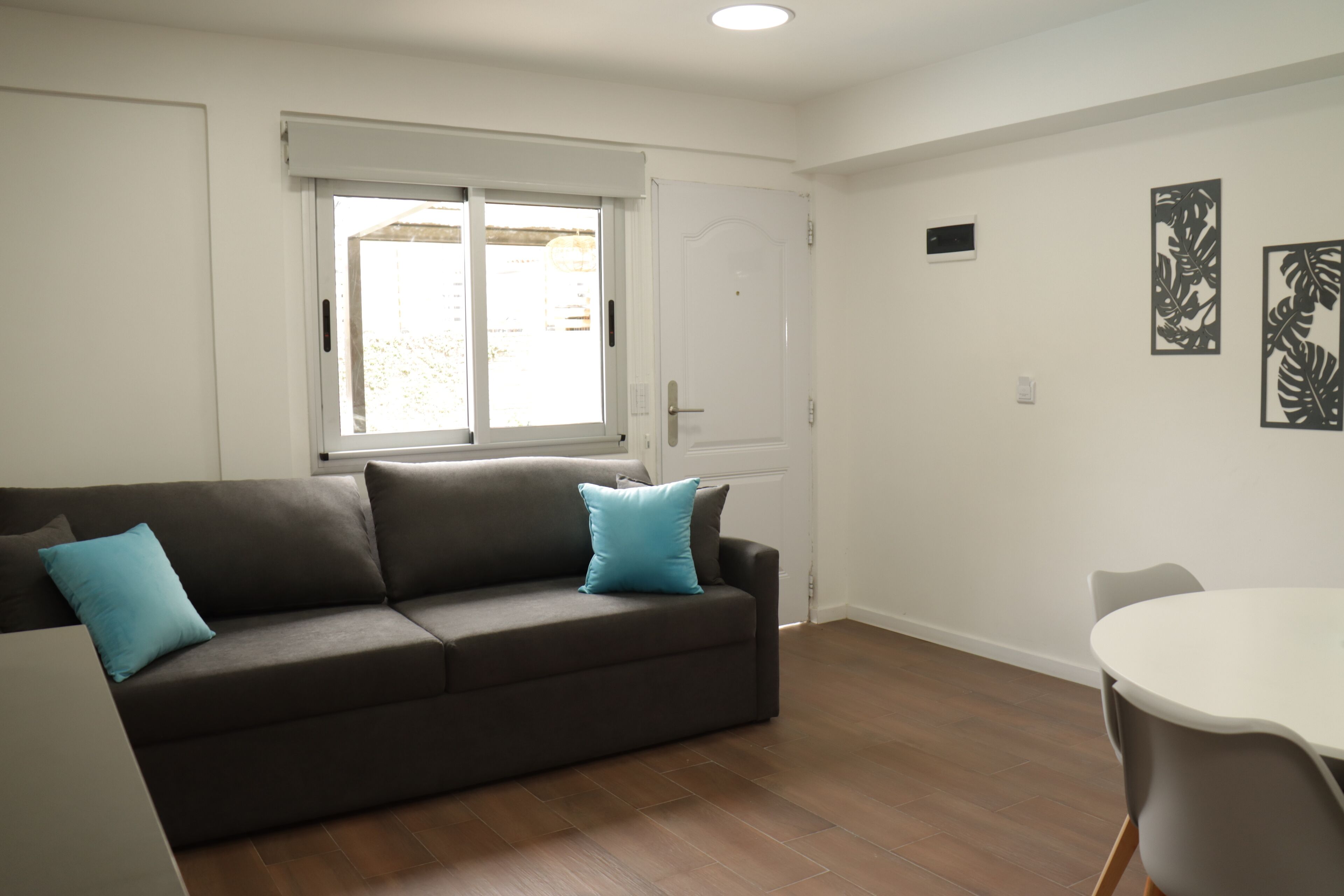 A living room with a brown sofa, two blue cushions, a white table, and a window with a view.