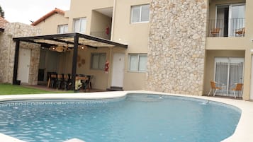A swimming pool with a stone wall, a covered patio, and a building with balconies.