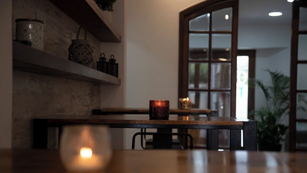 A wooden table with a candle in the foreground, a shelf with decorative items, and a window with a view of greenery. - ROZAS (Ezeiza)