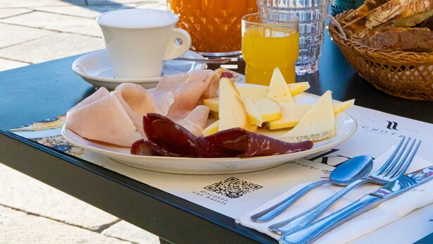 A table with a meal, including a pitcher of orange juice, a cup, and a plate with cheese and meats, set against a scenic mountain backdrop. - LVG Hotel Collection - Il Portico (Cannobio)