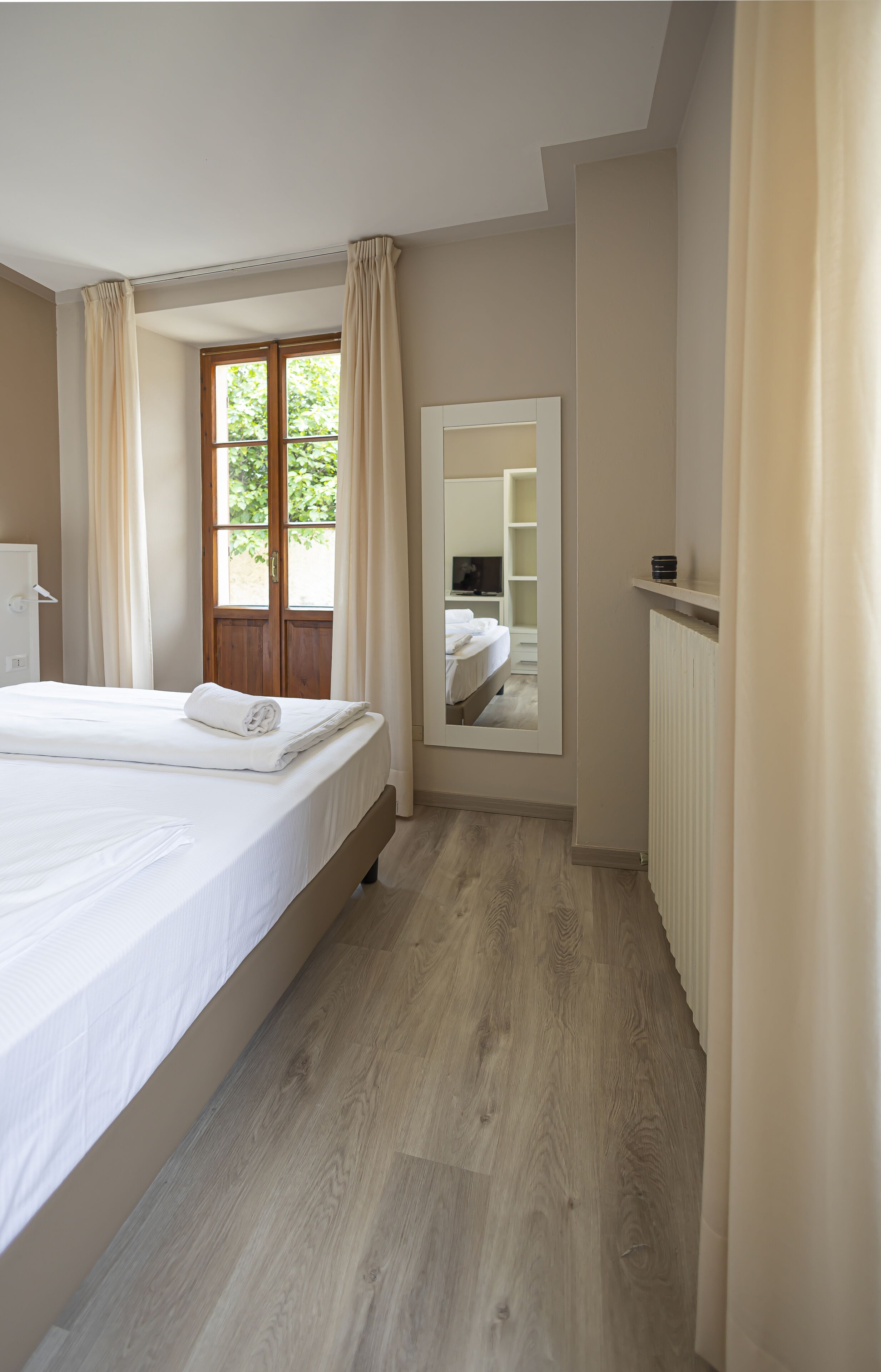 Un dormitorio con una cama grande, suelo de madera, una ventana con cortinas, un espejo y un radiador.