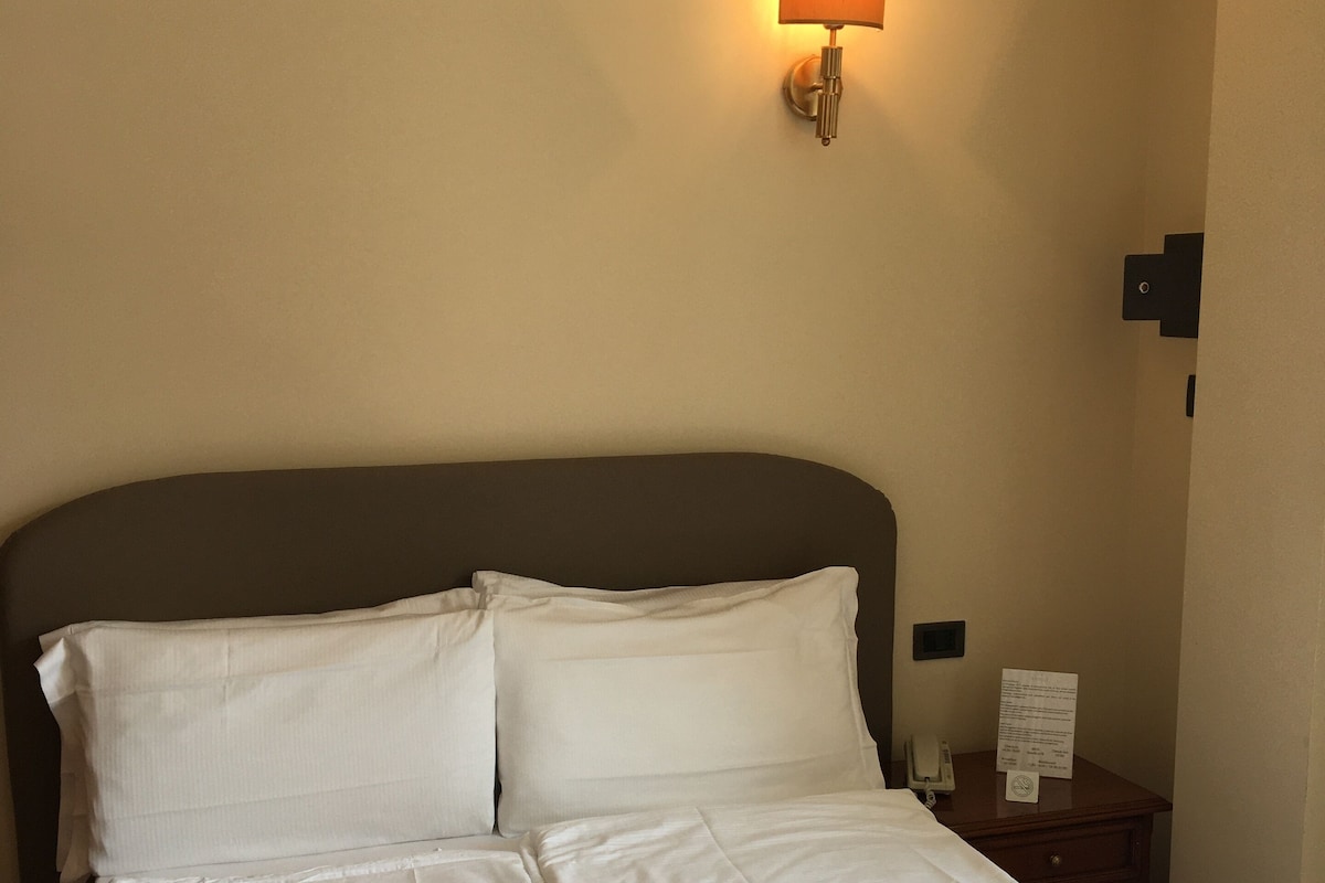 Una cama bien tendida con sábanas blancas y dos toallas enrolladas, una lámpara de pared y una mesita de noche pequeña con un teléfono y un periódico.