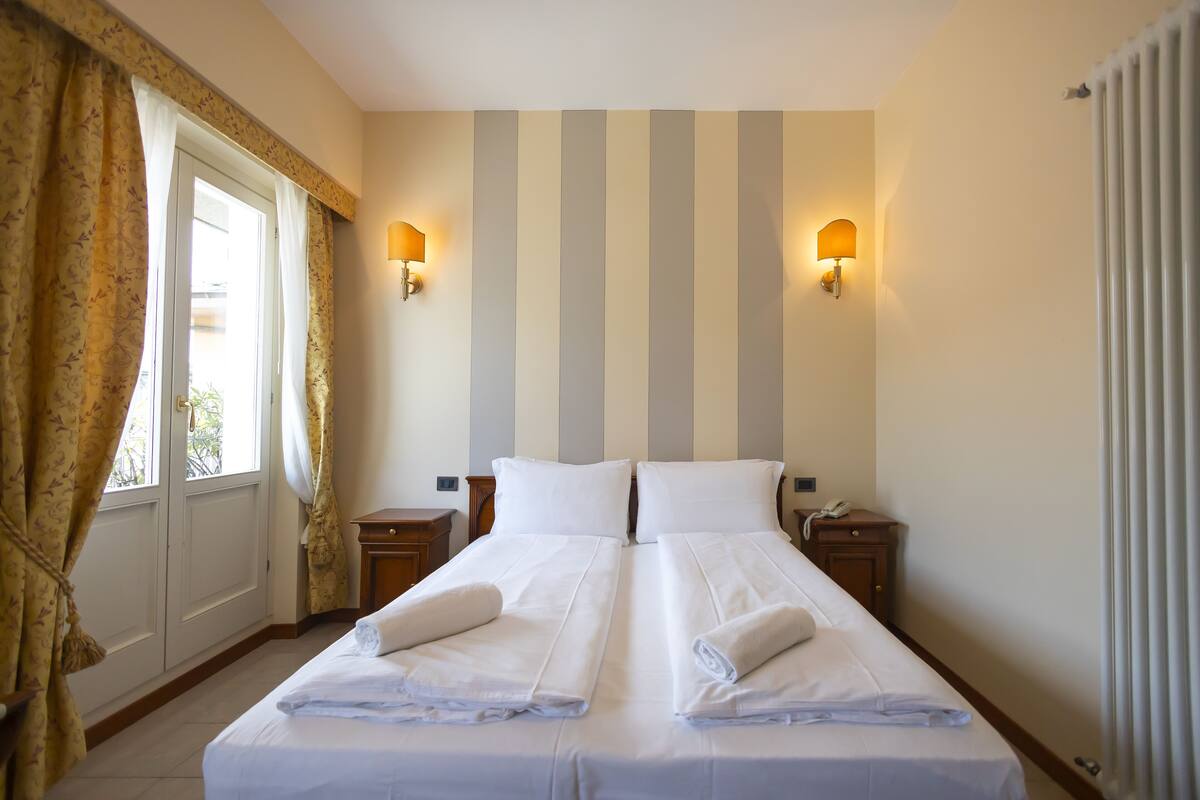 Un lit double avec du linge de lit et des serviettes blanches, deux tables de chevet, une fenêtre avec des rideaux et des motifs rayés sur les murs.
