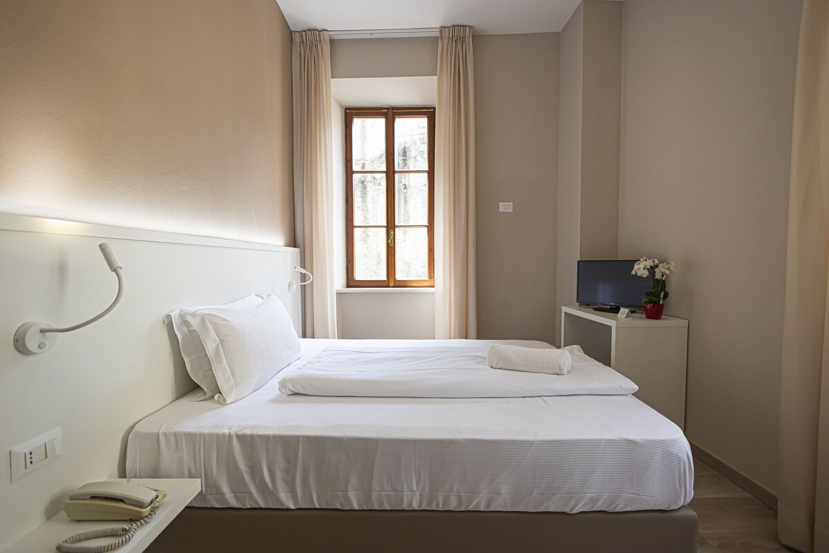 A hotel room with a bed, a bedside telephone, a TV, and a window with curtains.