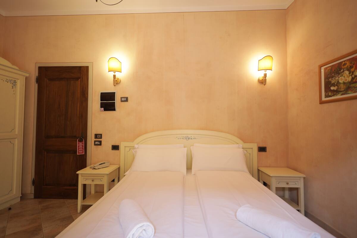 Une chambre d’hôtel avec une porte en bois, deux tables de chevet, un lit avec du linge de lit blanc et des lampes fixées au mur.