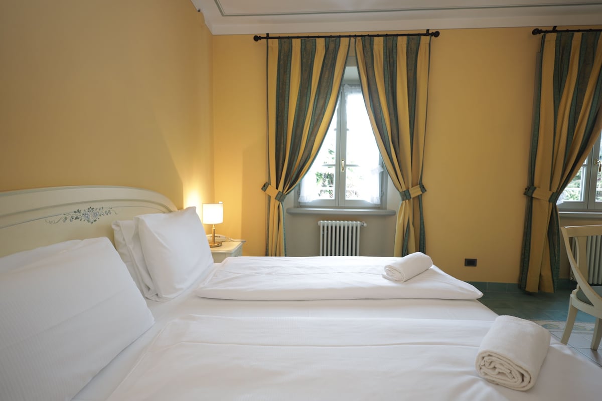Una cama bien hecha con sábanas blancas, una lámpara de noche y cortinas en una ventana.