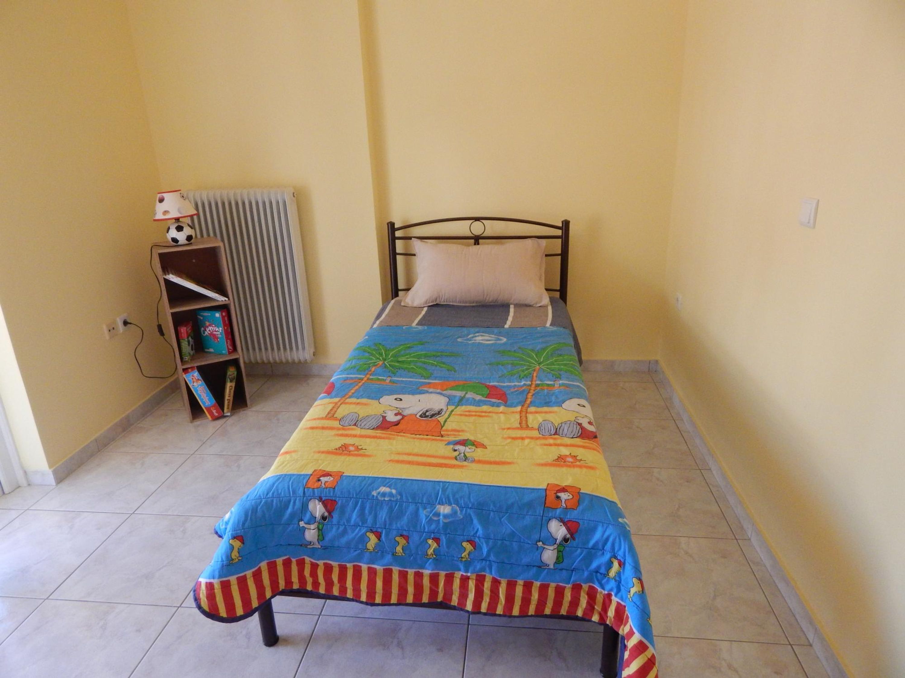 A small, single-bed room with a colorful bedspread, a wooden shelf with books, and a radiator.