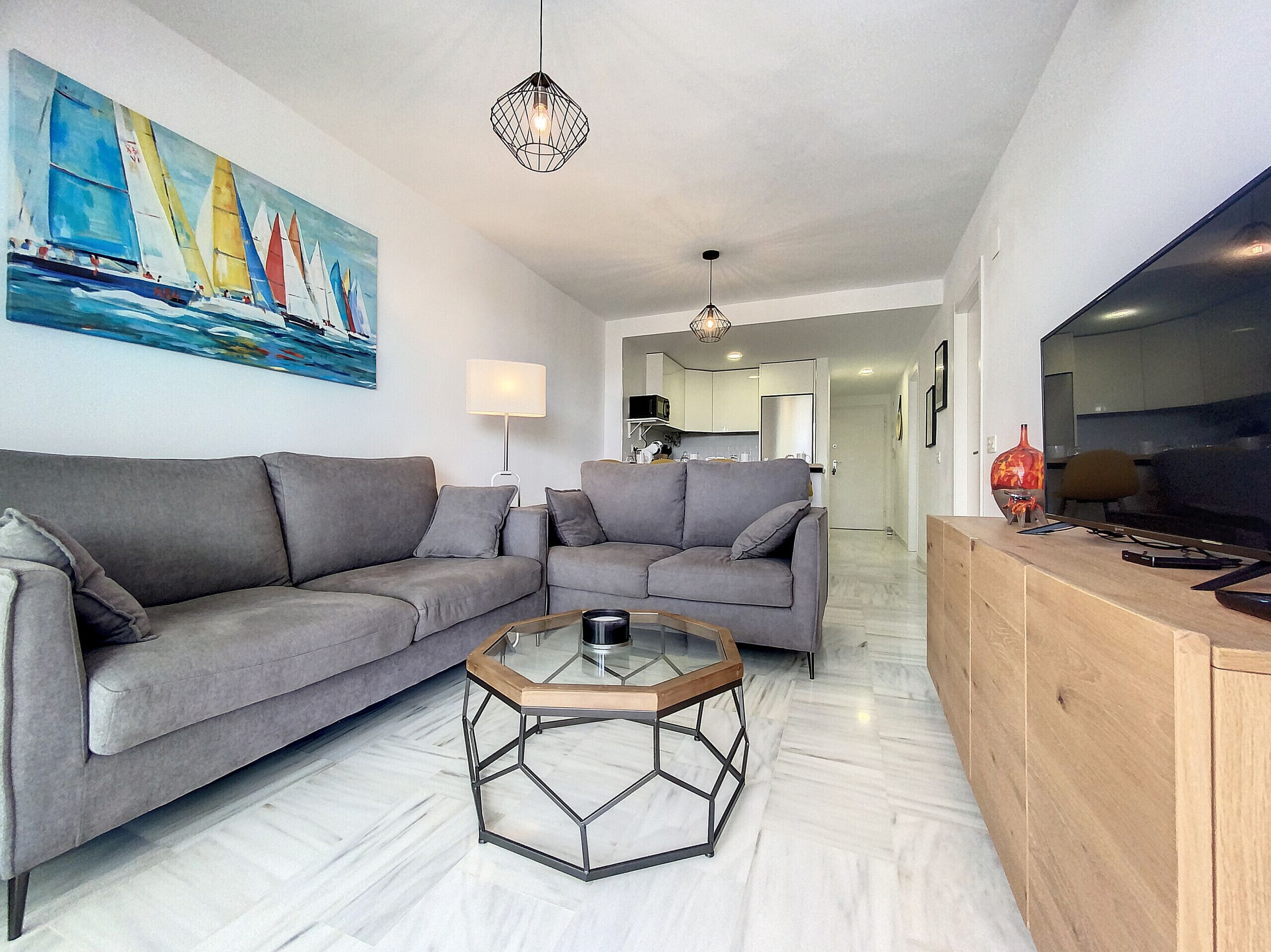 A modern living room with a grey sofa, a glass coffee table, and a painting of sailboats on the wall.
