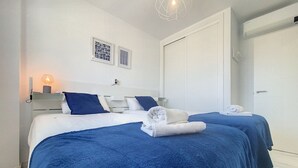 Ein Hotelzimmer mit zwei Betten, jeweils mit blauer Bettwäsche und weißen Handtüchern. Ein Nachttisch mit Lampe, ein Wandbild und eine Tür auf der rechten Seite sind ebenfalls vorhanden.