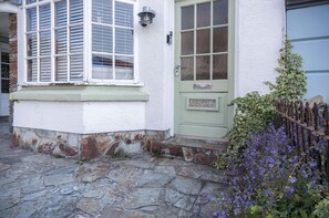 Een huis met een oprit van stenen, een groene deur met een naambordje, en een raam met luiken.