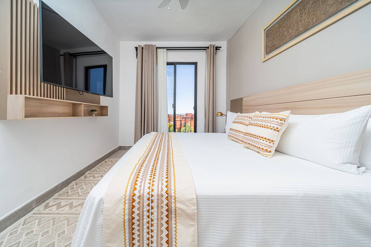 Ein Hotelzimmer mit Bett, Wandfernseher, einem Fenster mit Vorhängen und Blick auf Gebäude.