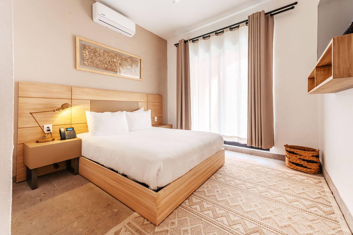 Ein Hotelzimmer mit einem Bettgestell aus Holz, einer Nachttischlampe, einem Telefon, einer Wandklimaanlage, einem Fenster mit Vorhängen und einem gewebten Korb.