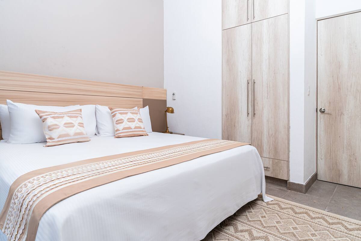 Ein ordentlich bezogenes Bett mit Holz Kopfteil, einer beigefarben-weißen Musterdecke und passenden Kissen. Rechts daneben befindet sich ein Holfschrank mit Griff und Tür.