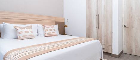 Cama bem arrumada com cabeceira de madeira, manta bege e branca estampada e almofadas combinando. Um guarda-roupa de madeira com puxador e porta à direita.