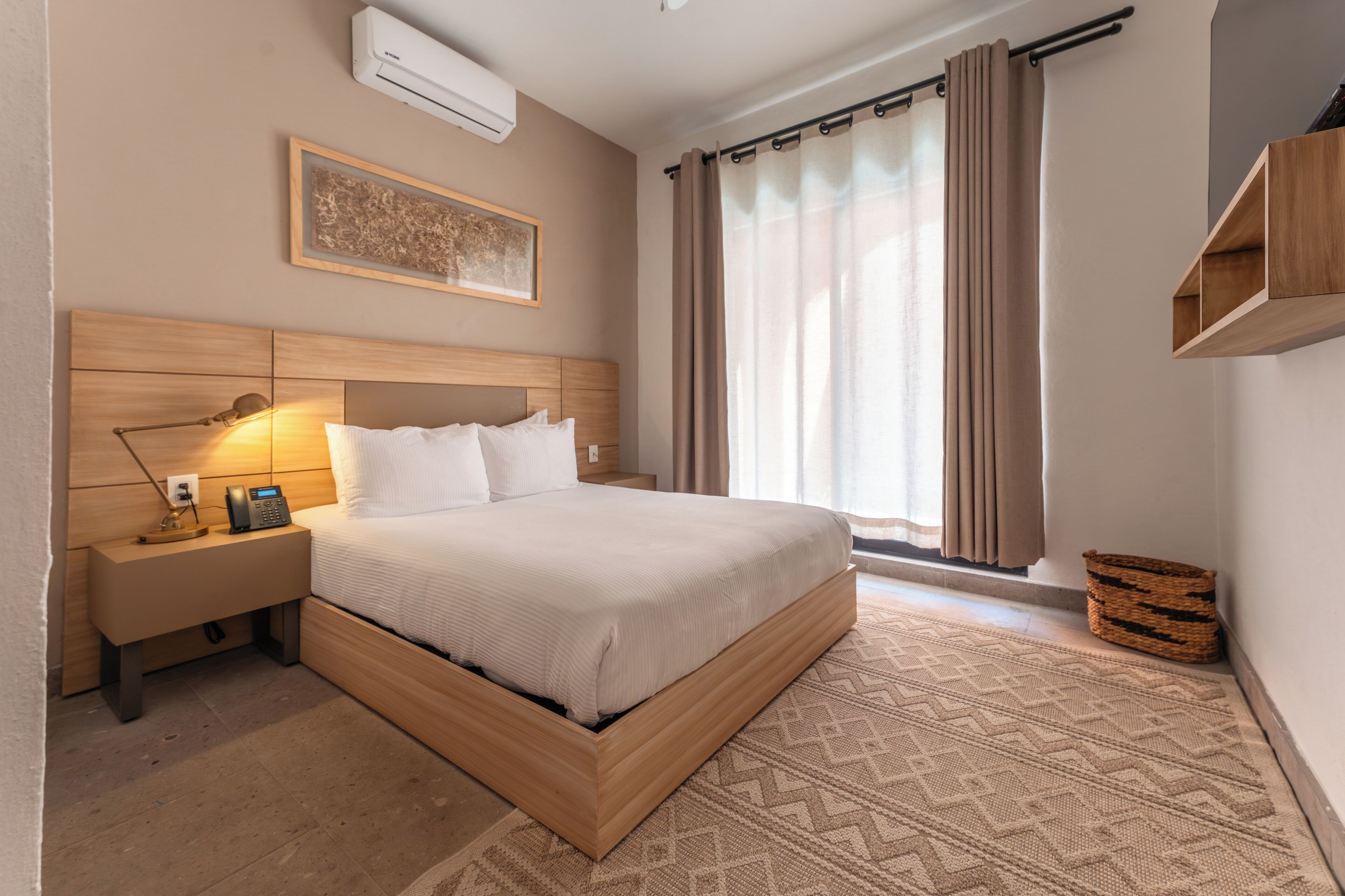 A neatly arranged bedroom with a wooden bed, bedside table with a lamp, a wall-mounted air conditioner, and a window with curtains.