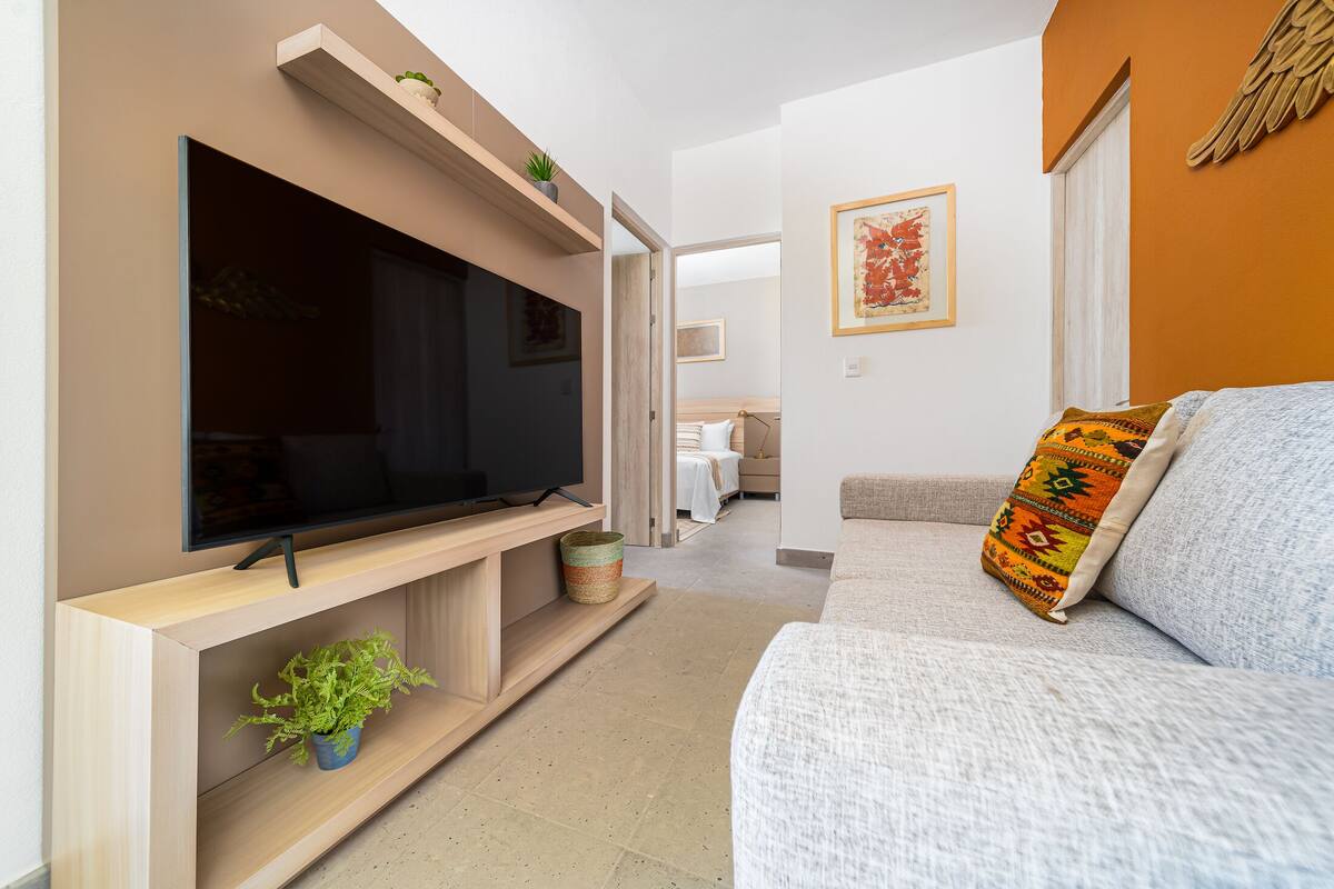 Ein modernes Wohnzimmer mit einem Flachbildfernseher auf einem Holzständer, einem grauen Sofa mit gemusterten Kissen und einer kleinen Pflanze auf dem Boden.