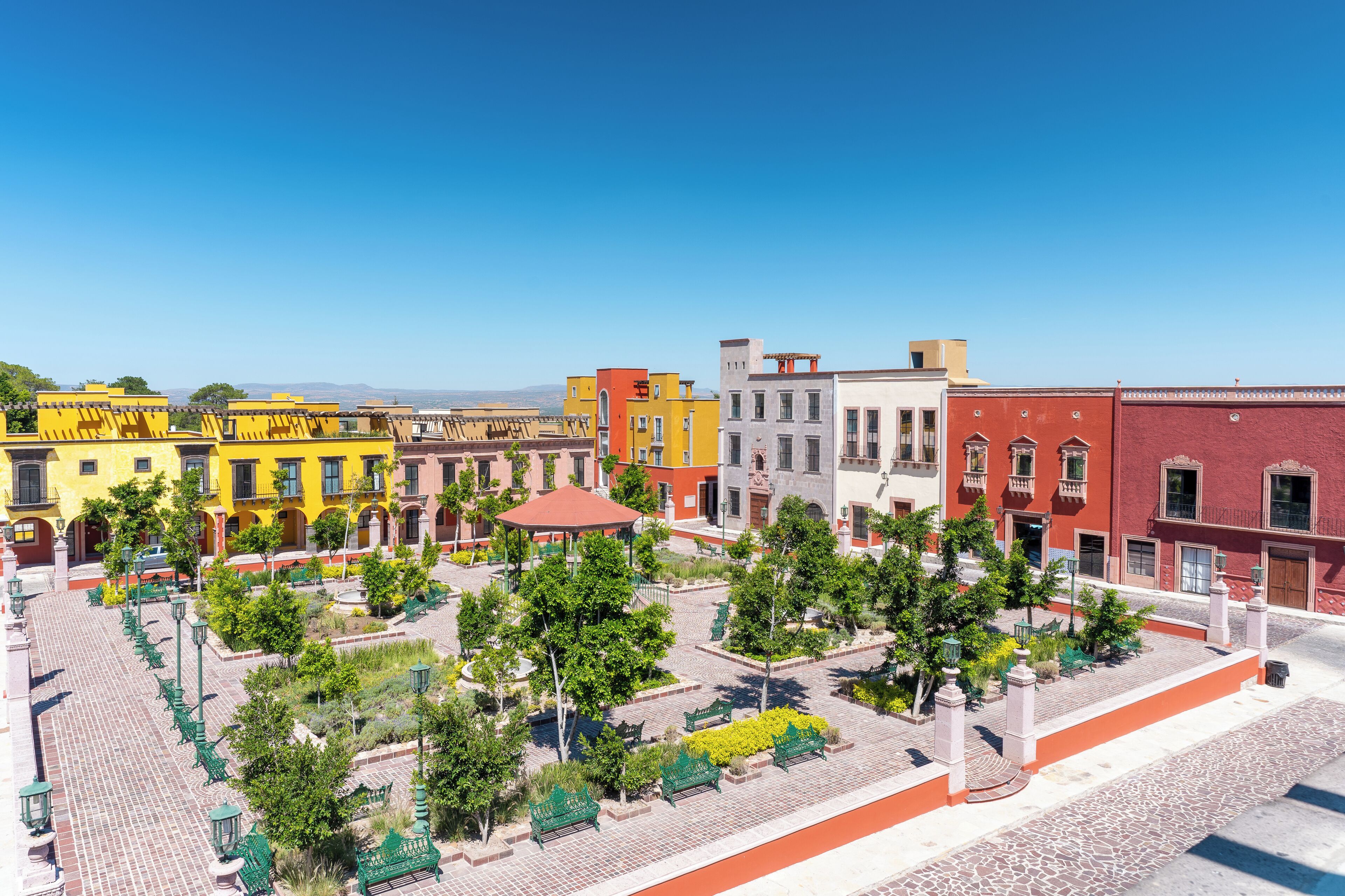 A plaza with colorful buildings, green benches, and trees.