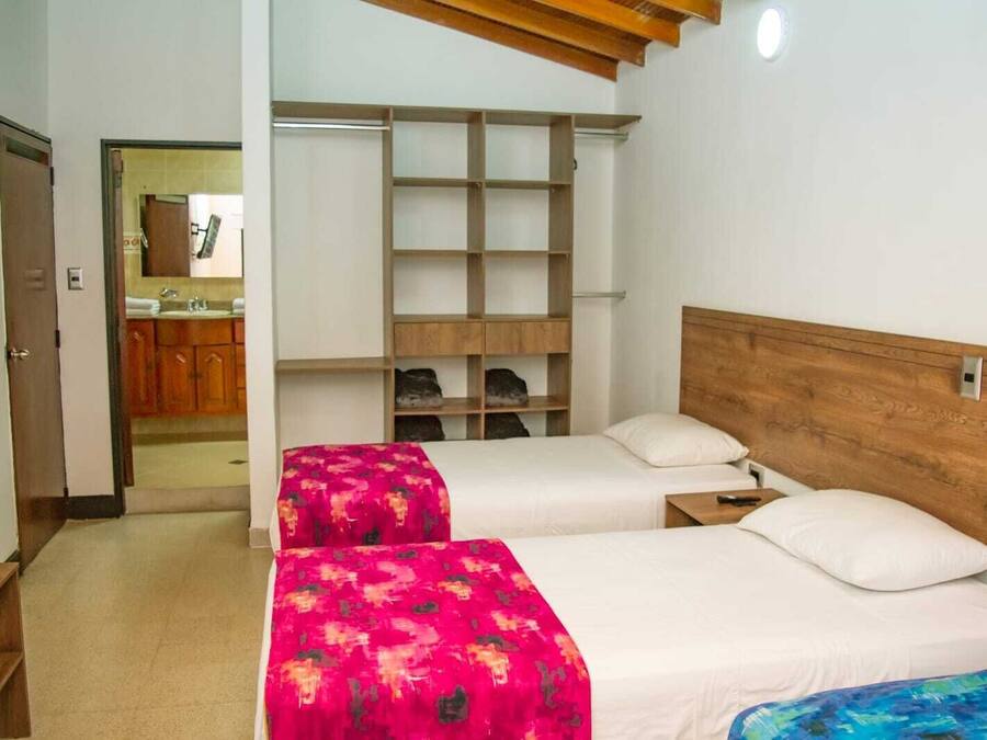 A hotel room with two beds, a wooden headboard, a built-in shelf unit, and a view of the kitchen area.