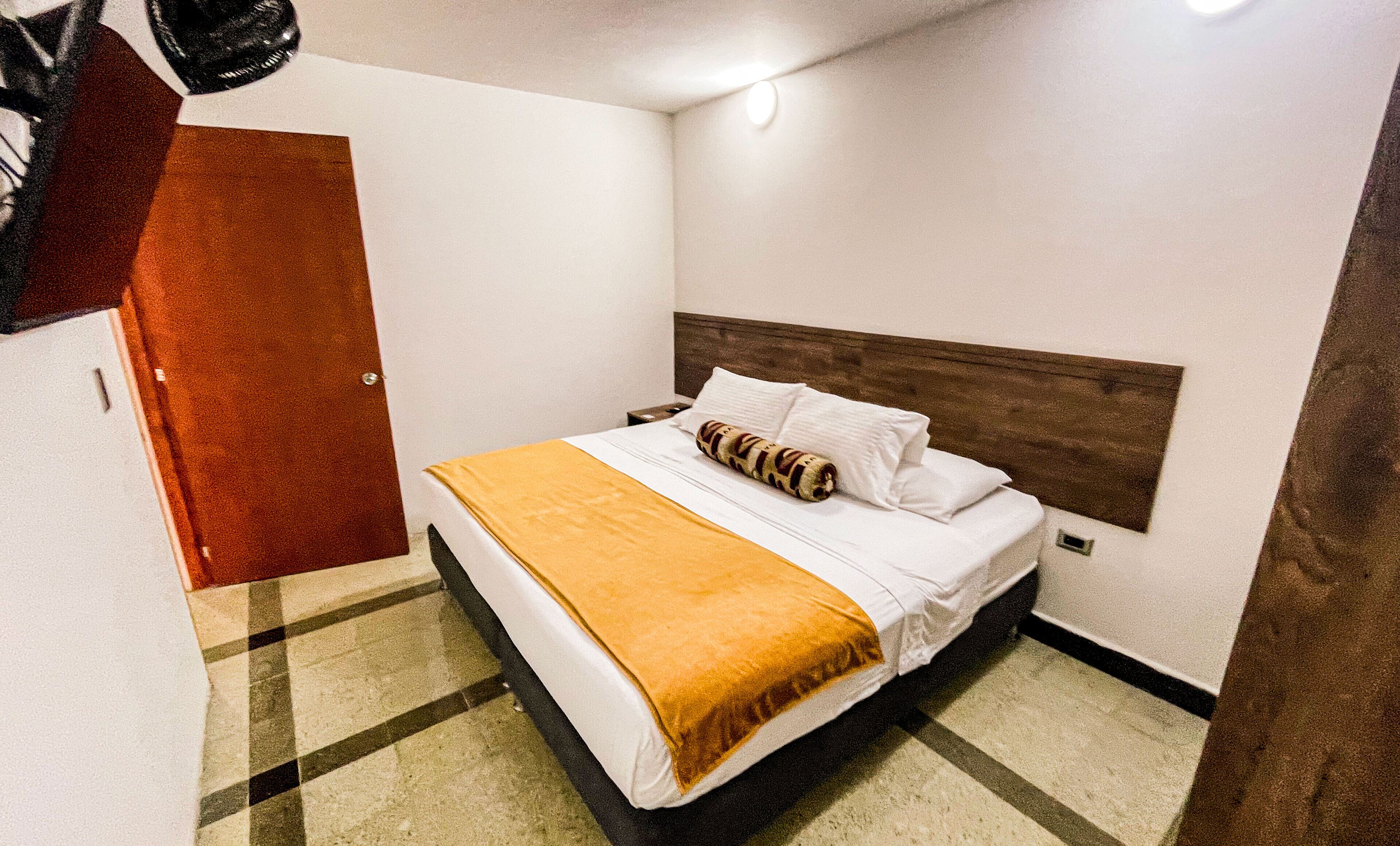 Een hotelkamer met een bed, nachtkastje, houten hoofdeinde en een geruit vloerkleed.