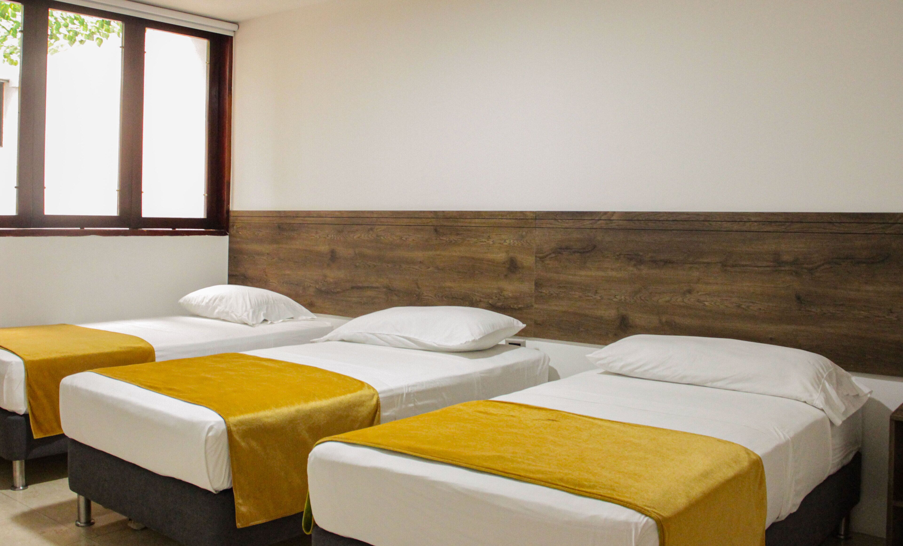 Una habitación con tres camas individuales alineadas junto a un cabecero de madera. Cada cama tiene un colchón blanco y una colcha amarilla.