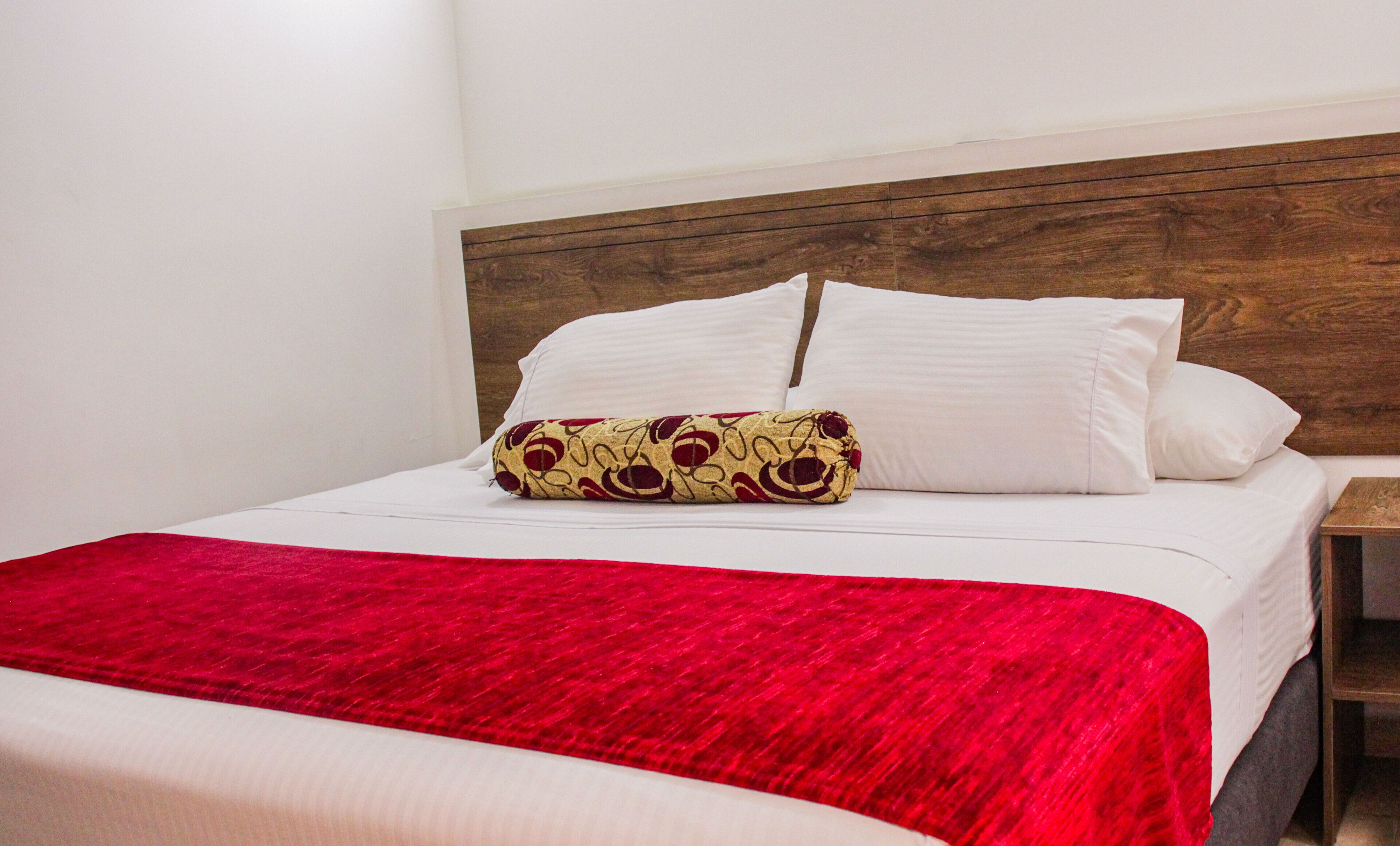 A neatly made bed with white linens, a red blanket, and a patterned pillow.