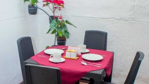 A table set with white plates, cups, and a small cake on a red tablecloth, surrounded by black chairs, with a potted plant on the wall. - Hotel Boutique Estadio Medellin (Medellín)