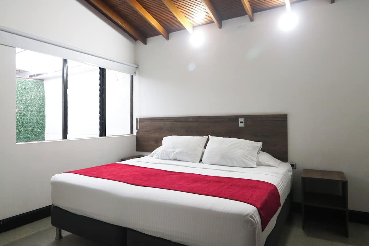 Ein Schlafzimmer mit einem Bett, einer roten Decke, einem Holzkopfteil, einem Nachttisch und einem Fenster mit Blick auf Grünflächen.
