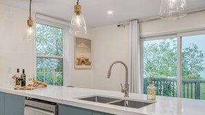 Une cuisine moderne avec un plan de travail blanc, un évier en acier inoxydable et un lave-vaisselle. On y trouve des luminaires suspendus et une fenêtre donnant sur un paysage verdoyant.