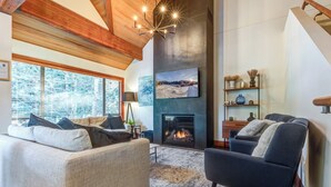A cozy living room with a fireplace, a large window with blinds, and a modern chandelier.