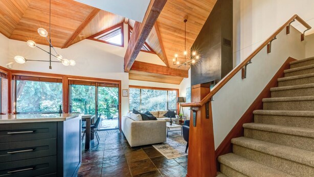 A spacious living area with a wooden staircase, a kitchen with dark cabinetry, and large windows offering a view of the outdoors.