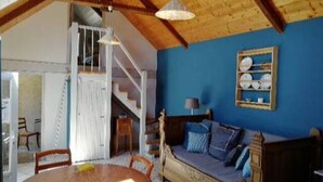 Une pièce chaleureuse avec un plafond en bois, un mur bleu, un canapé avec des coussins, une table à manger avec des chaises et un escalier menant à un autre niveau.