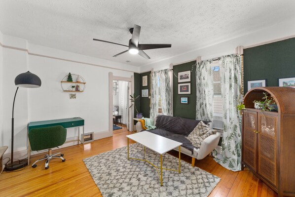 A living room with a desk, a sofa, a coffee table, a floor lamp, and a ceiling fan.