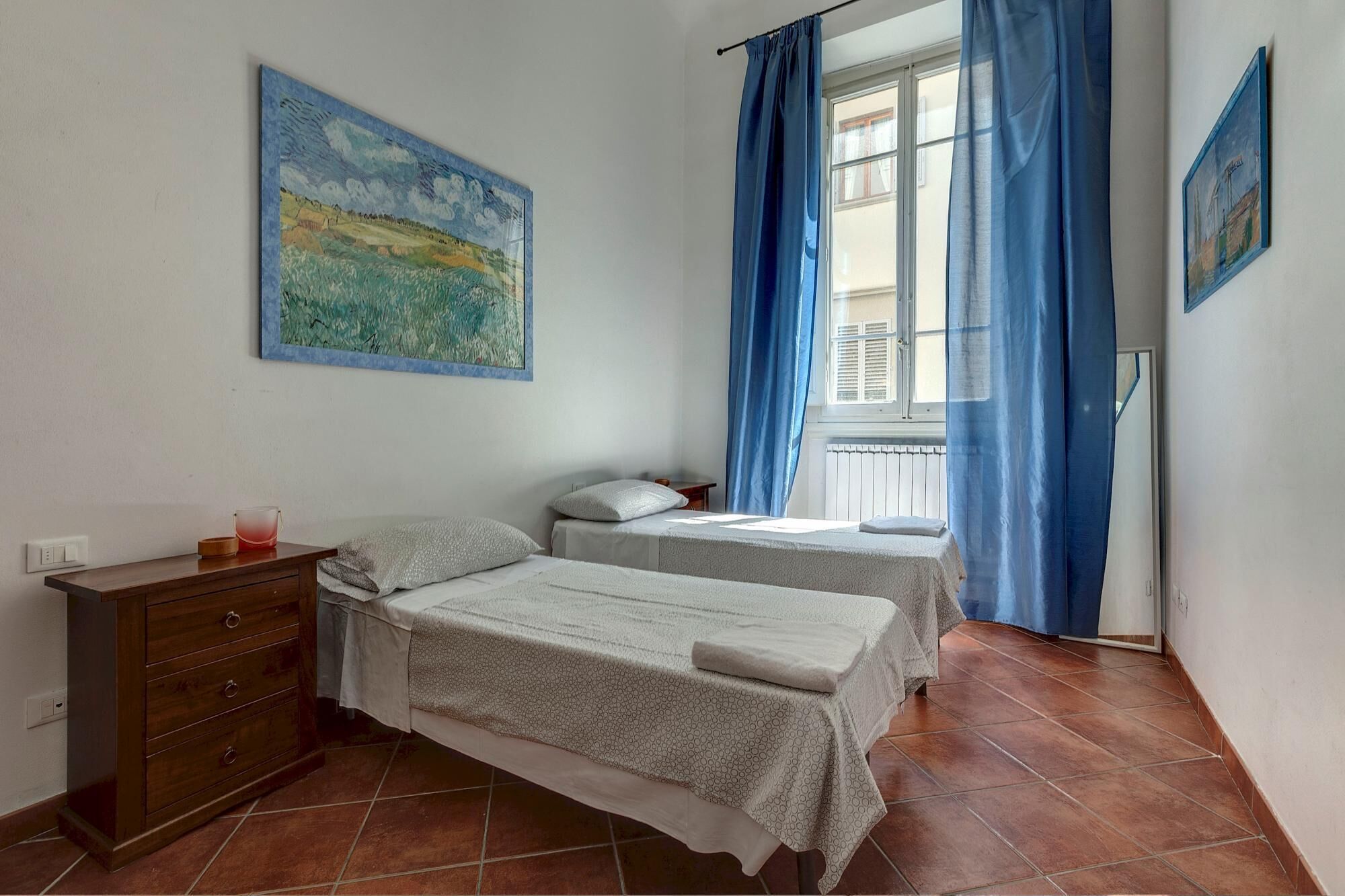 A bedroom with two beds, a wooden dresser, and a window with blue curtains.