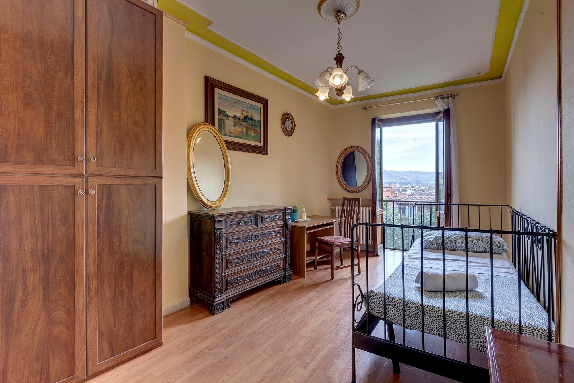A room with a wooden cabinet, a dresser, a desk, a chair, a bed with a view, and a balcony railing.