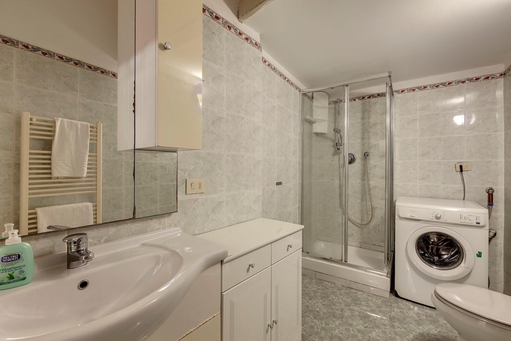 A bathroom with a sink, mirror, towel rack, shower, and washing machine.
