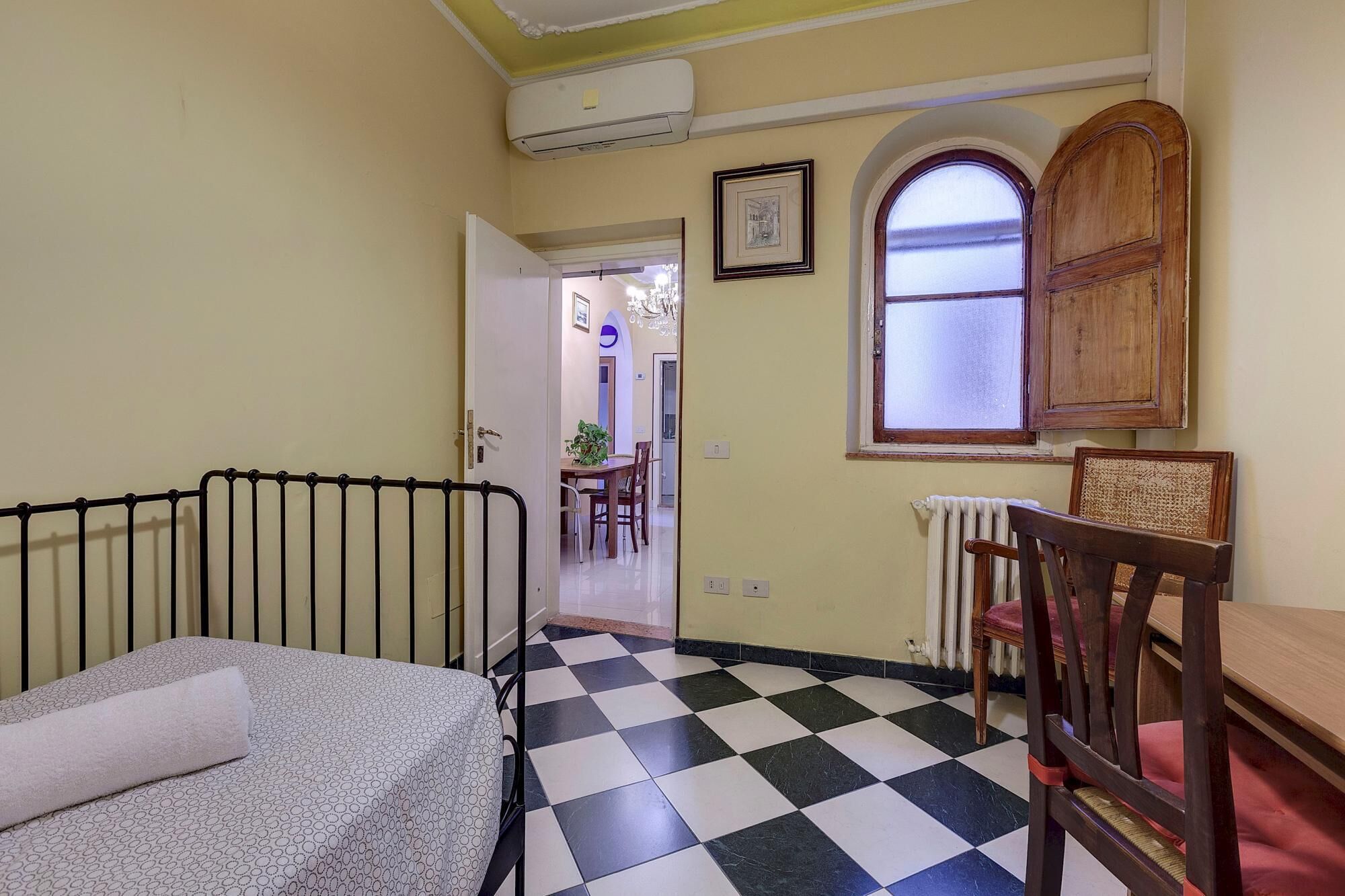 A room with a checkered floor, a bed with a patterned cover, a wooden chair, a radiator, and a framed picture on the wall.
