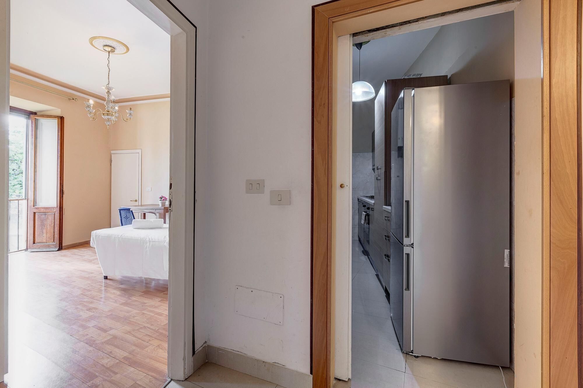 A hotel room with a wooden floor, a bed, a chandelier, and a modern stainless steel refrigerator.