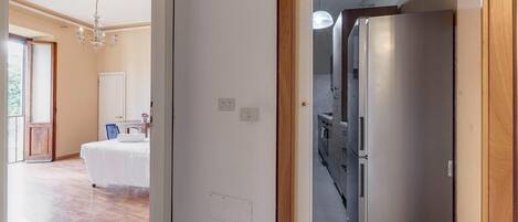 A hotel room with a wooden floor, a bed, a chandelier, and a modern stainless steel refrigerator.