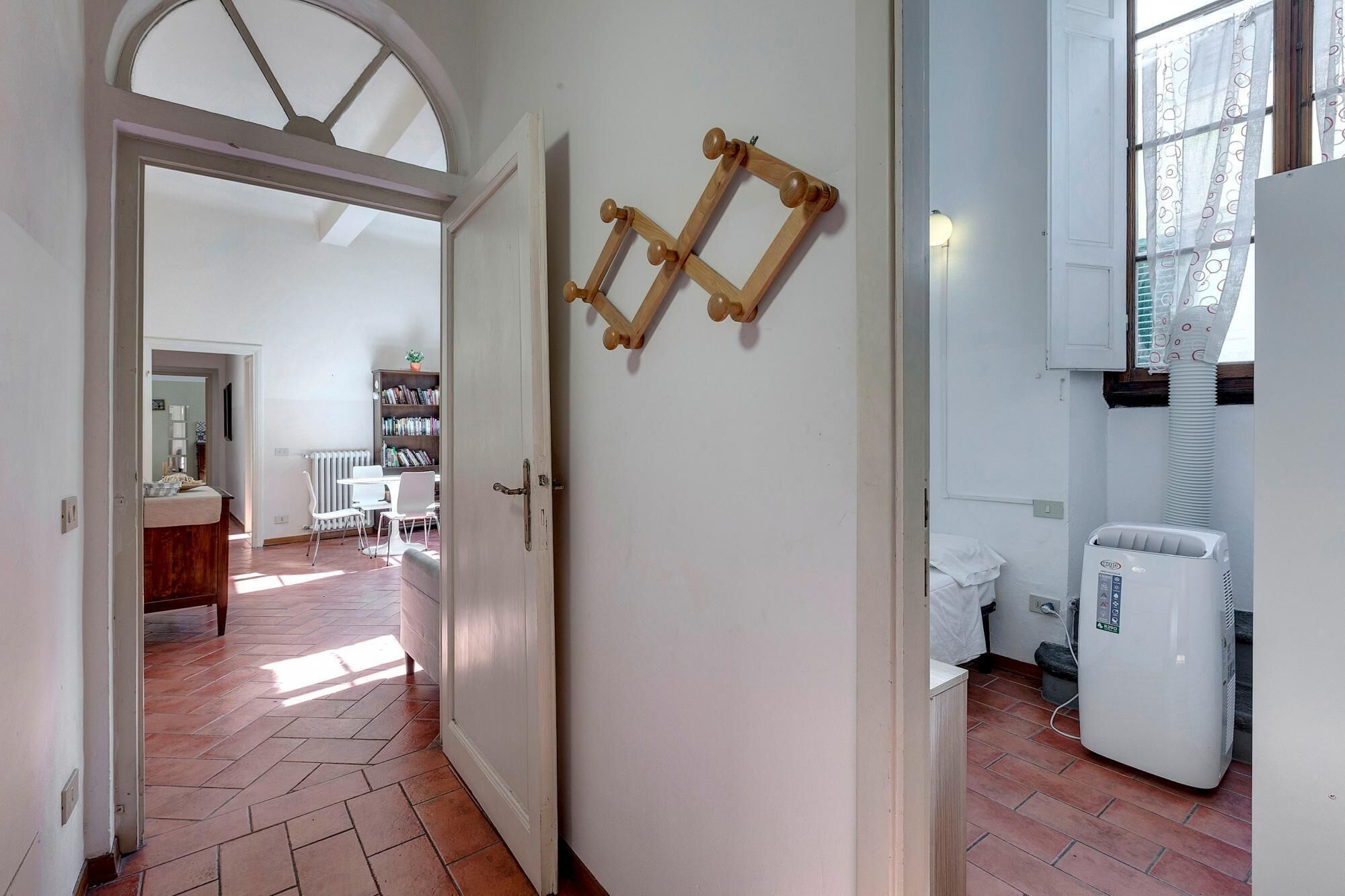 Um corredor com chão de azulejos que leva a uma sala de jantar, com mesa, cadeiras, estante e radiador.