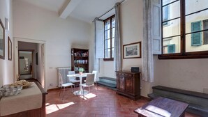 A room with a tiled floor, a round table with chairs, a bookshelf, a radiator, and a window with curtains. - Neri 23 in Firenze With 3 Bedrooms and 2 Bathrooms (Florence)