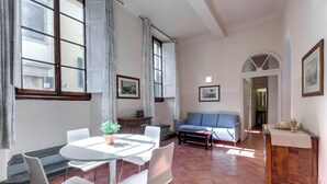 A spacious living area with a dining table, white chairs, a blue sofa, and a coffee table. There are large windows with curtains, a staircase, and a hallway leading to another room. - Neri 23 in Firenze With 3 Bedrooms and 2 Bathrooms (Florence)